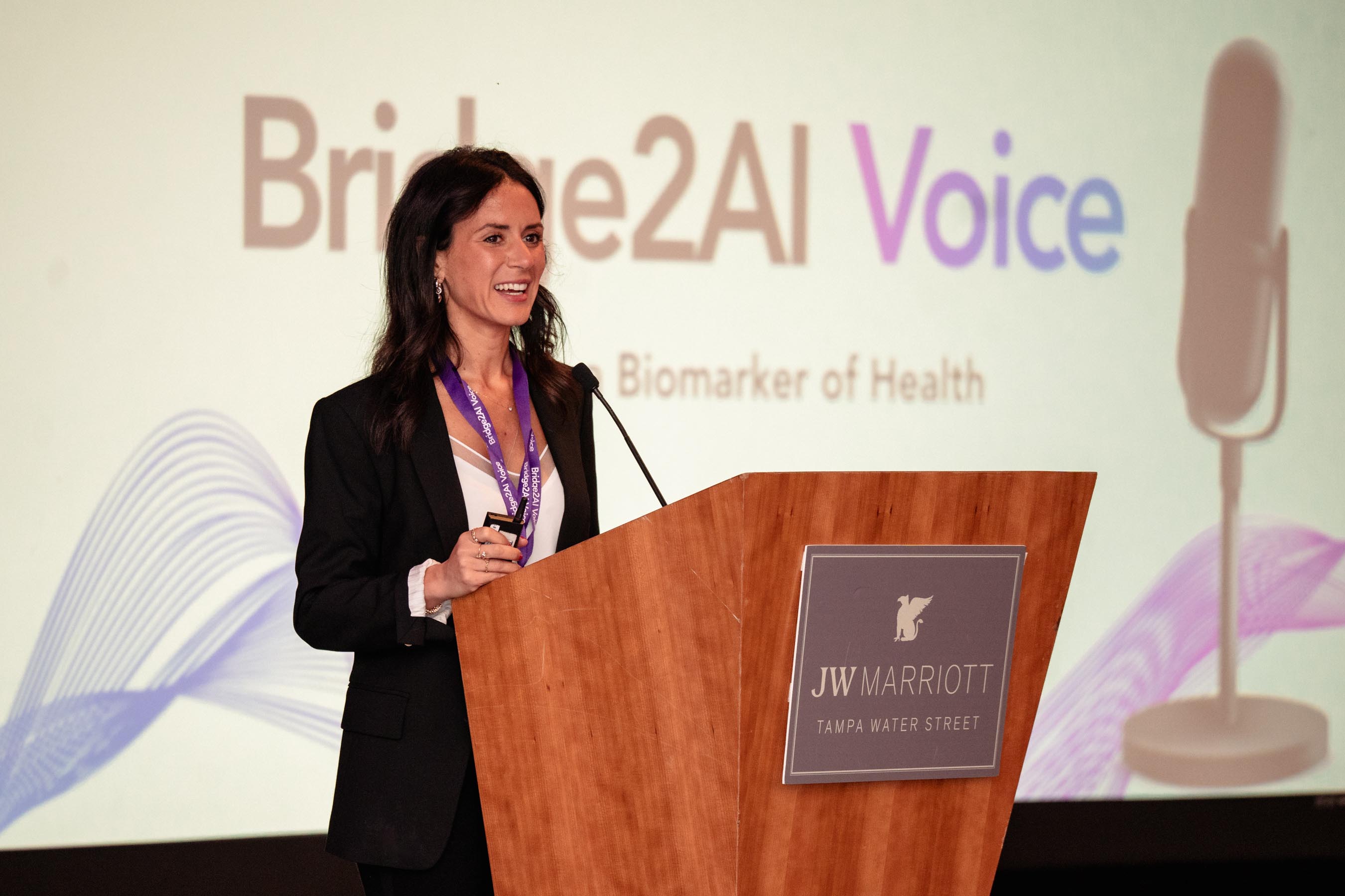 Dr. Yael_Bensoussan speaking at a podium with a black blazer and white shirt on. A screen behind her reads: Bridge 2AI Voice