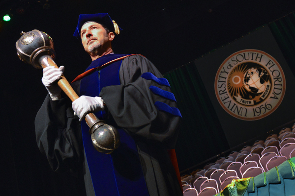 USF Health Faculty member holding specially designed mace wearing a black and blue graduation cap and gown. He is standing in front of chairs and a logo that reads: University of South Florida 1956, Truth and Wisdom