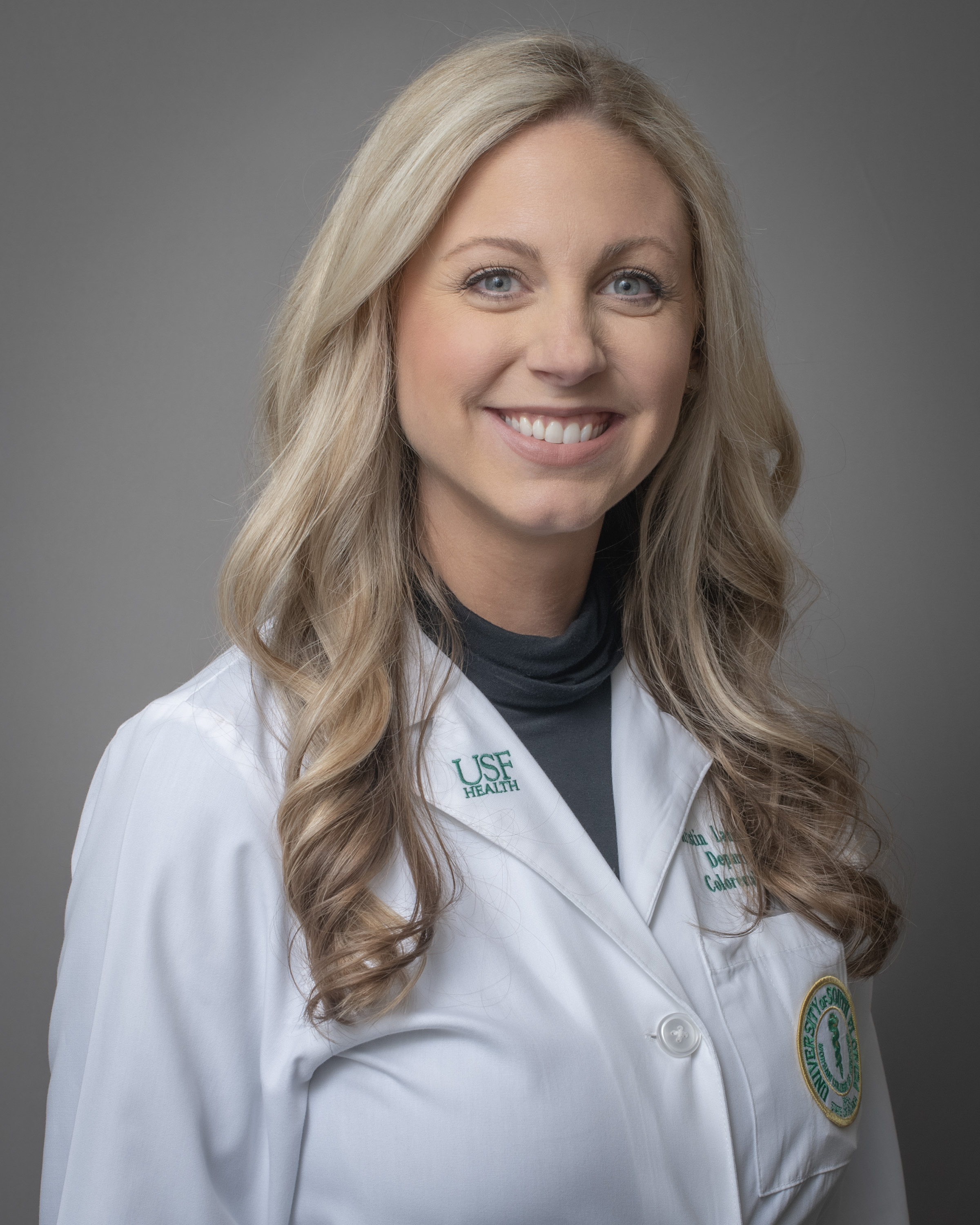 Kristin Launikitis smiling wearing a medical white coat with long blonde curled hair