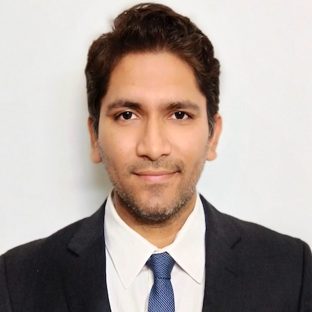 A man with dark hair and brown eyes smiles wearing a black suit with a blue tie