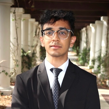 A man with dark hair and glasses smiles in a black suite with a black tie