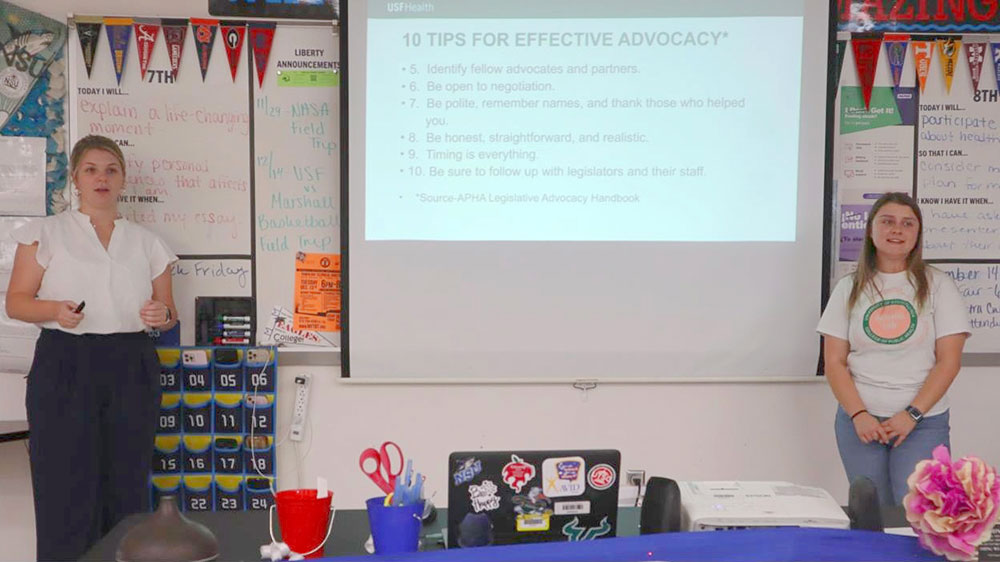Two people standing on either side of a whiteboard in a school classroom, with colorful educational materials and posters visible on the walls.