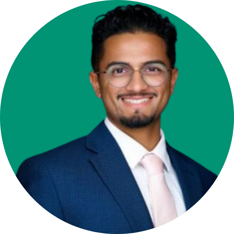 A headshot of Kishan Patel wearing a navy blue suit jacket, white dress shirt, and baby pink tie. He is smiling and posed in front of a green backdrop.