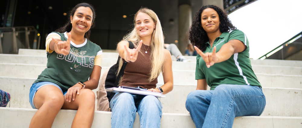 Three students holding up the "Go Bulls!" hand symbol as they sit on a staircase outdoors on campus.