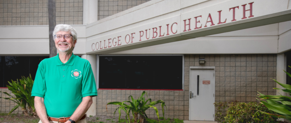 Dr. Sten Vermund standing in front of the entrance to the College of Public Health. He is smiling and wearing a green shirt.