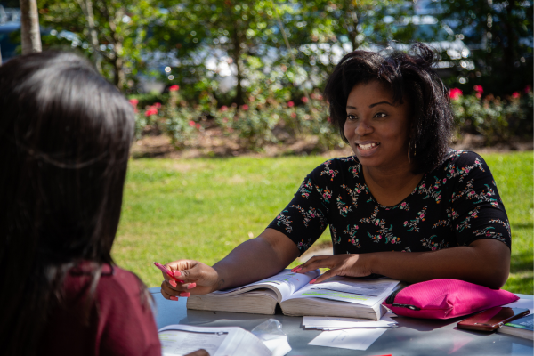 A woman is seated at a table, reading a textbook, surrounded by a warm and inviting outdoor environment.