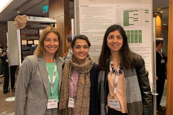 Three women stand side by side in front of a colorful poster, smiling and engaging with the camera.