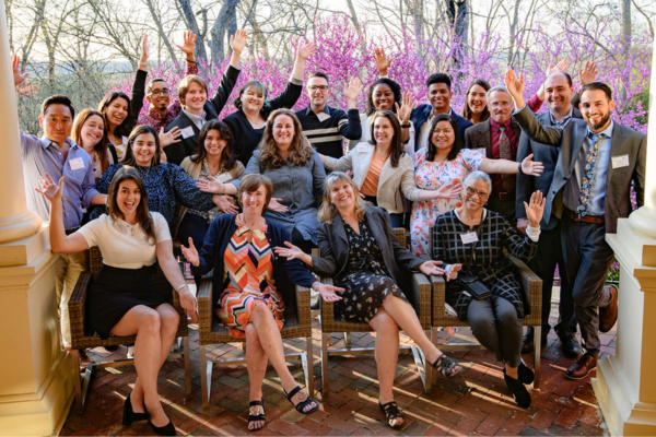 A group of people smiling and posing together on a porch, surrounded by greenery and sunlight.