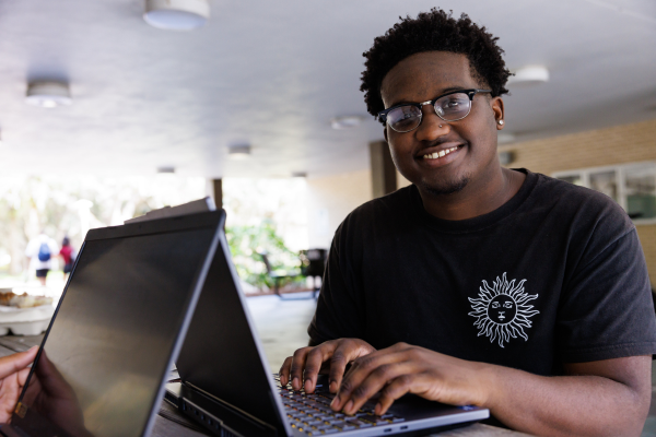 A student posing for a photo, smiling while typing on their laptop.