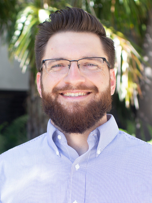 A headshot of Dr. Matthew Valente in an outdoor setting with a palm tree behind him.He is smiling and wearing a blue button-up shirt.