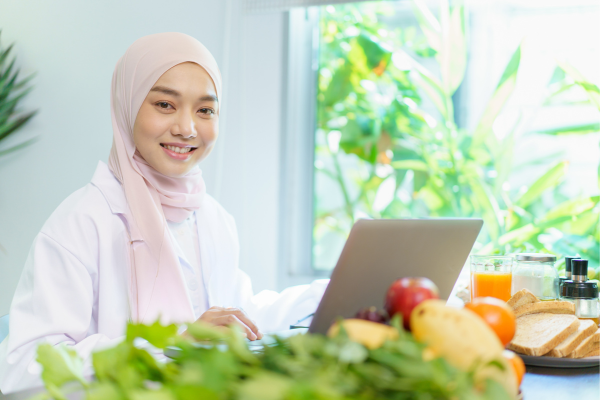 A student in a hijab, smiling and working on her laptop.