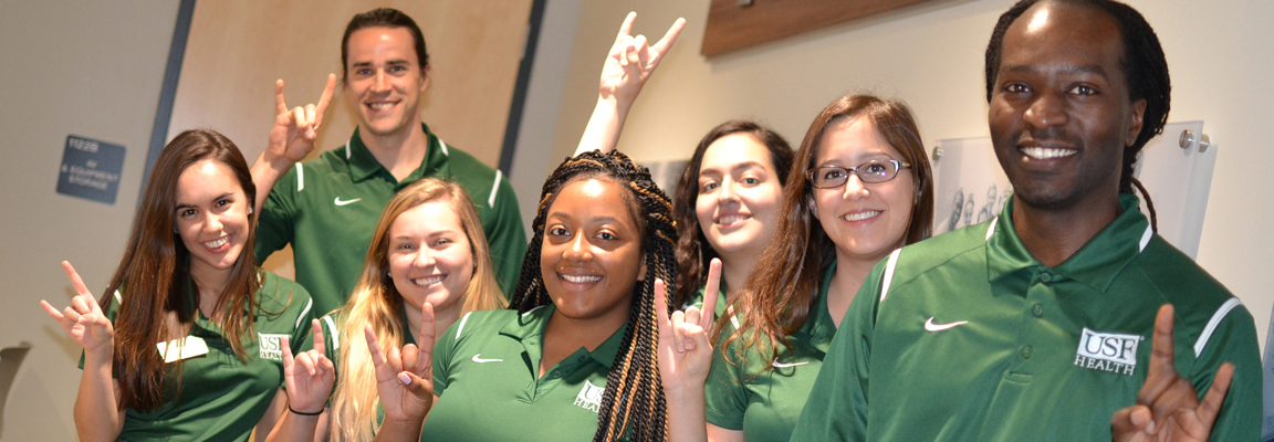 Group of men in women in green USF health polo shirts making the bull sign with their lifted hands.