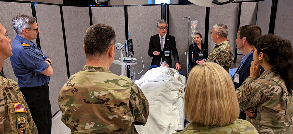 A group of military personnel stands around a hospital bed, discussing a patient’s condition.  