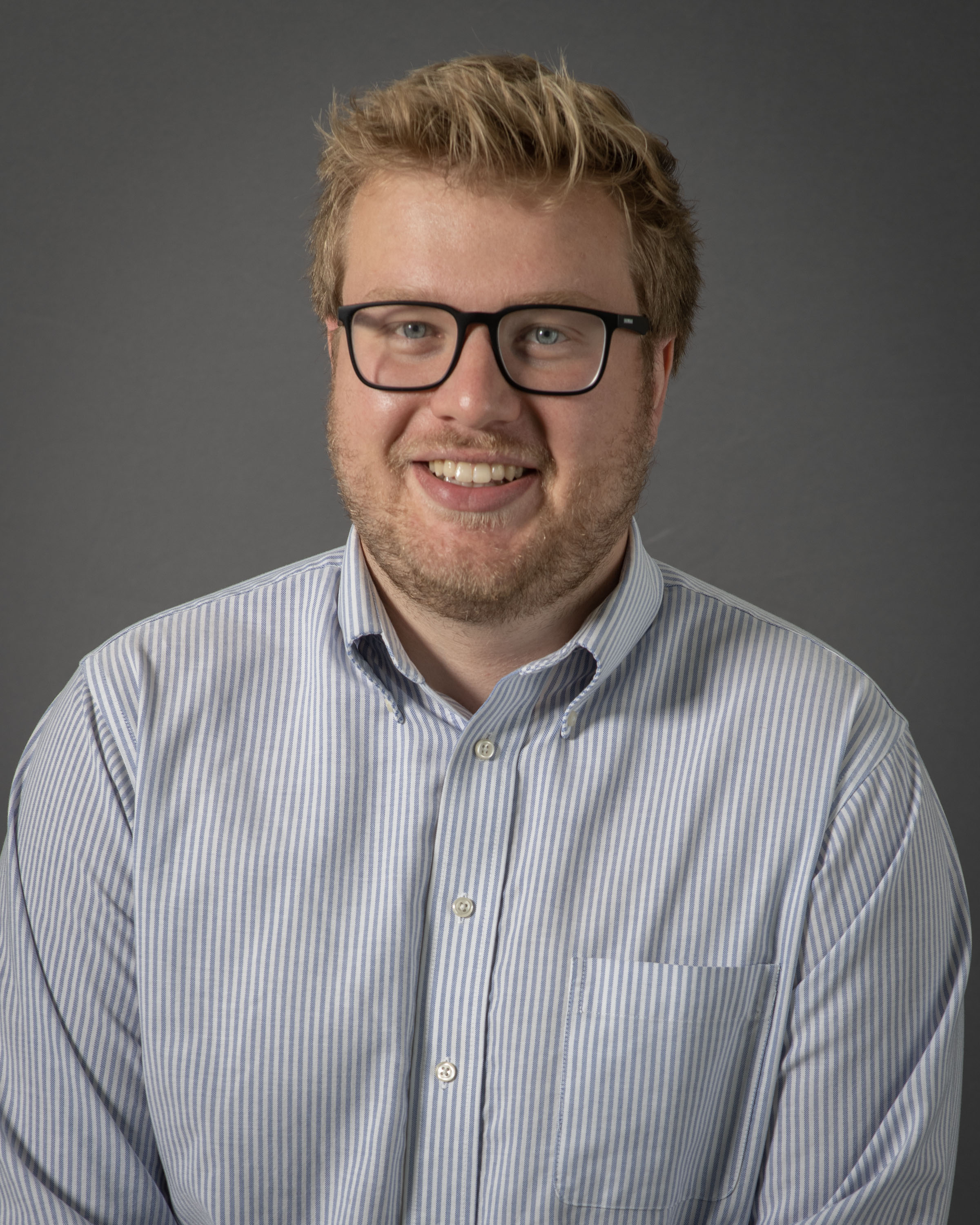 Van Ediger smiling wearing a blue and white striped dress shirt, grey background