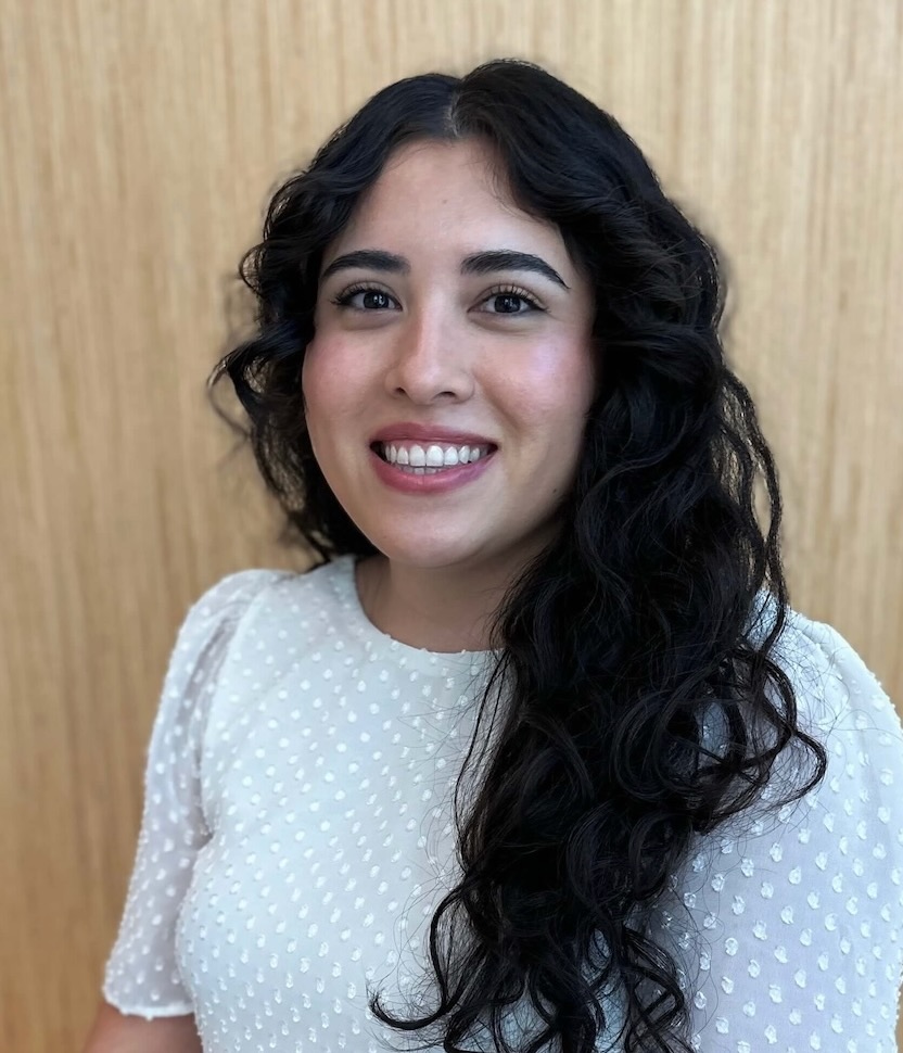 Katherine Vargas smiling wearing a white blouse with white polka dots in front of a light wood background