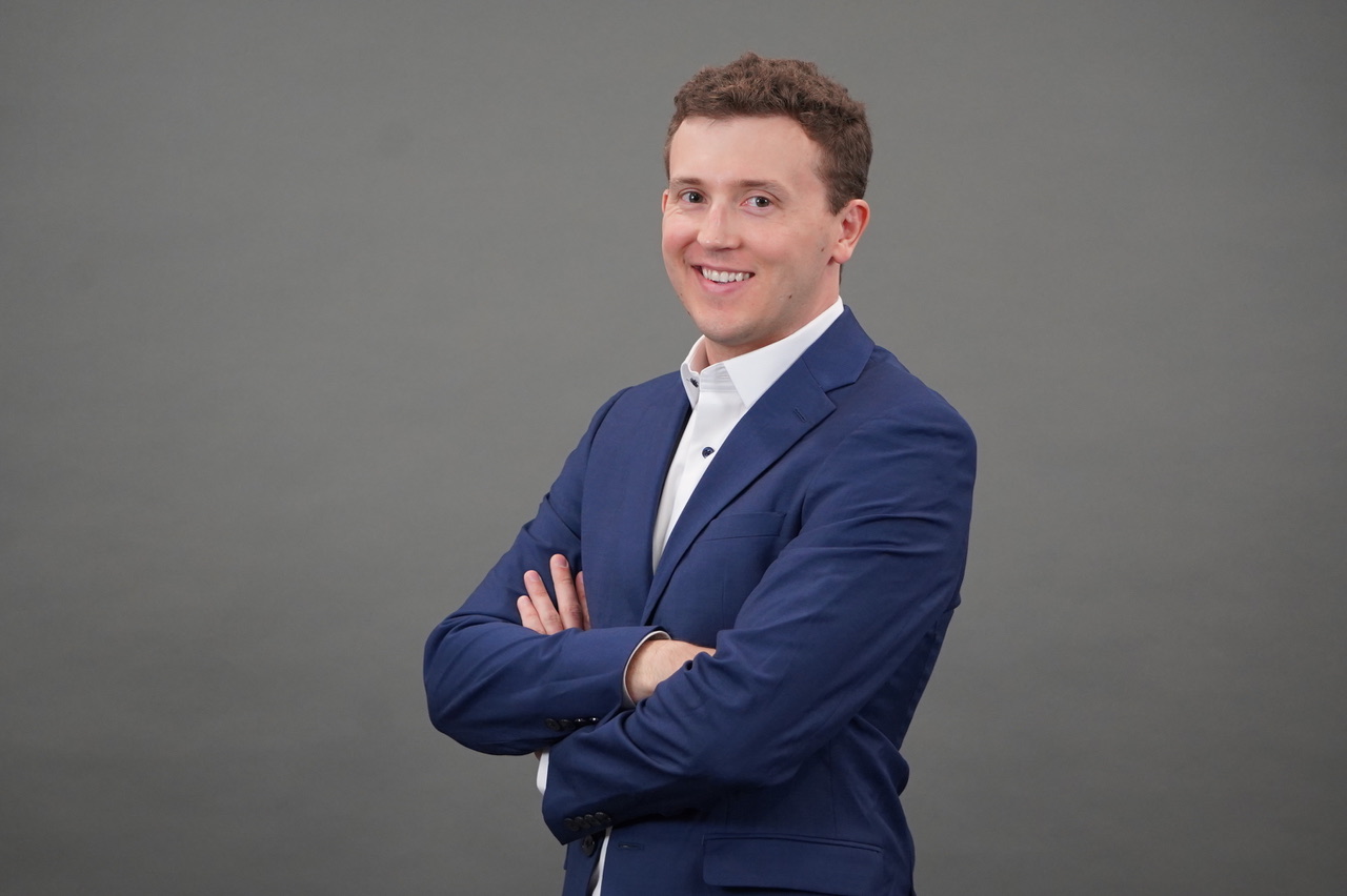Mitch Hansen smiling wearing a blue suit and white collard shirt crossing his arms
