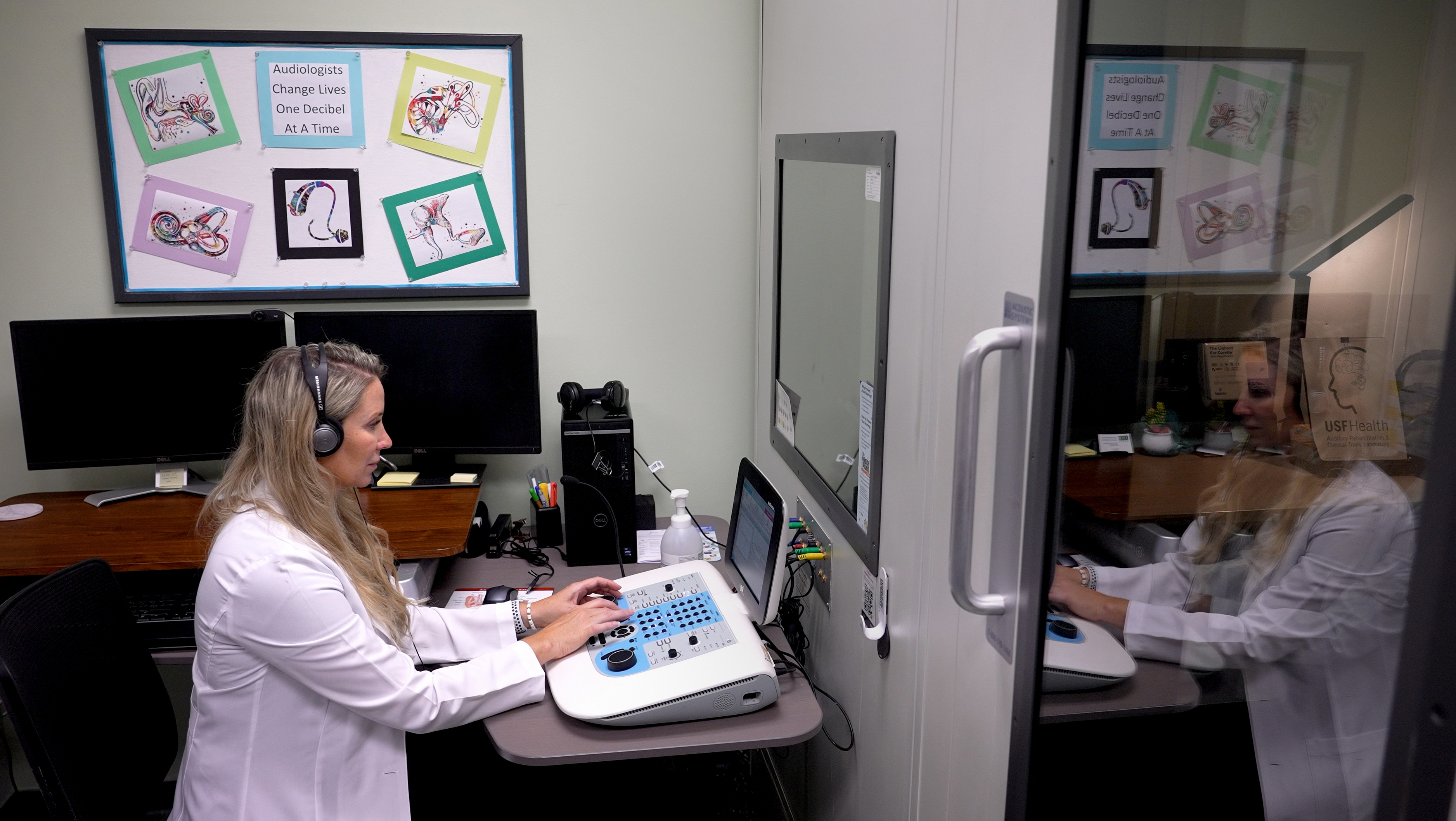 Dr. Victoria Sanchez sitting wearing a medical white coat and headphones conducting hearing research looking through a glass window