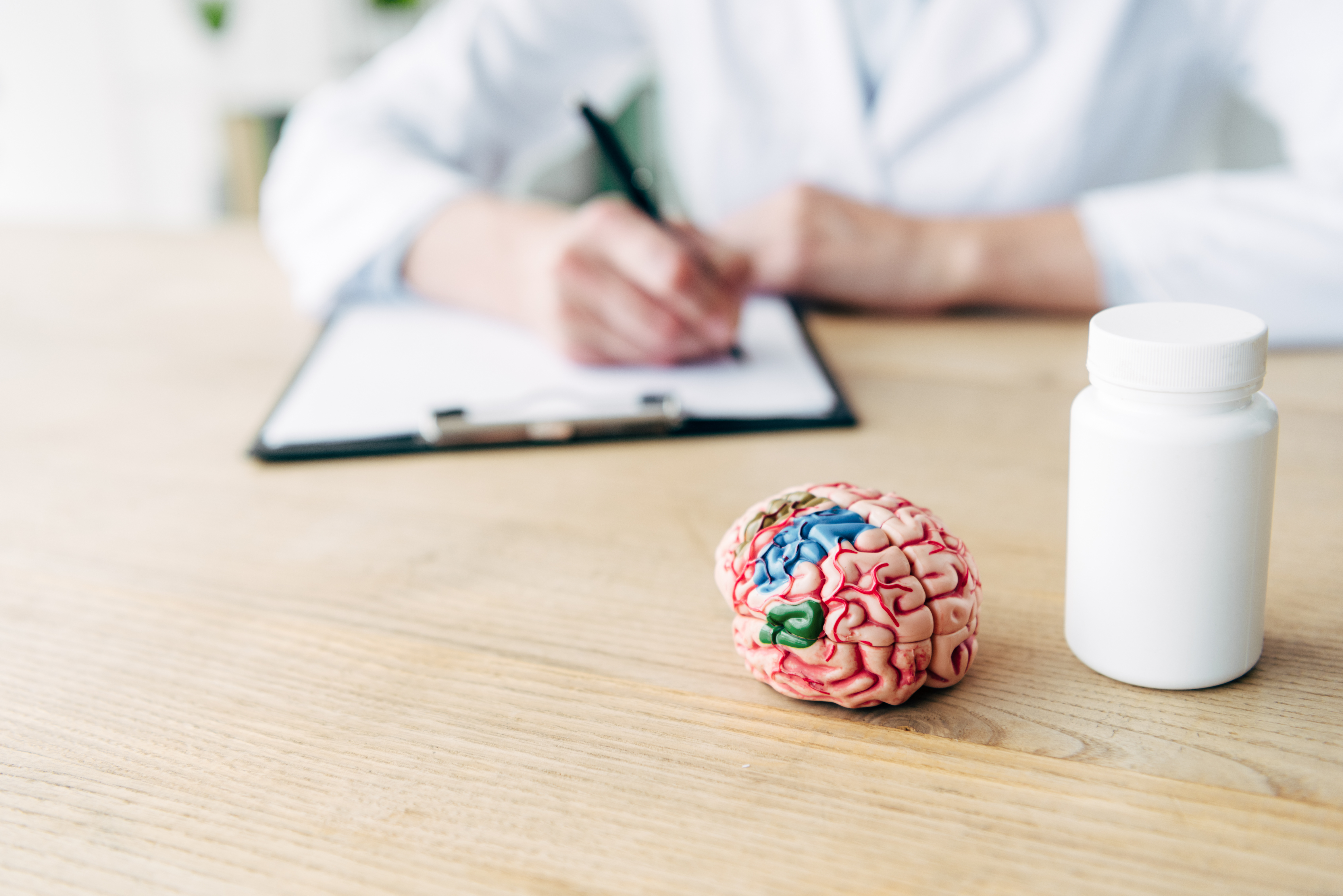 "A medical doctor in a white coat is writing on a piece of paper with a black pen at a table. Nearby, there is a model of a brain and a bottle of medicine, representing treatments for age-related diseases."