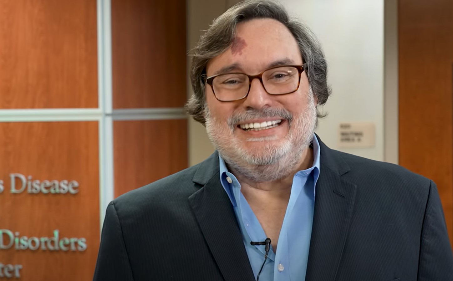 Dr. Robert Hauser smiling in a blue dress shirt and black blazer in front of a sign that reads: USF Health Parkinson’s Disease & Movement Disorders