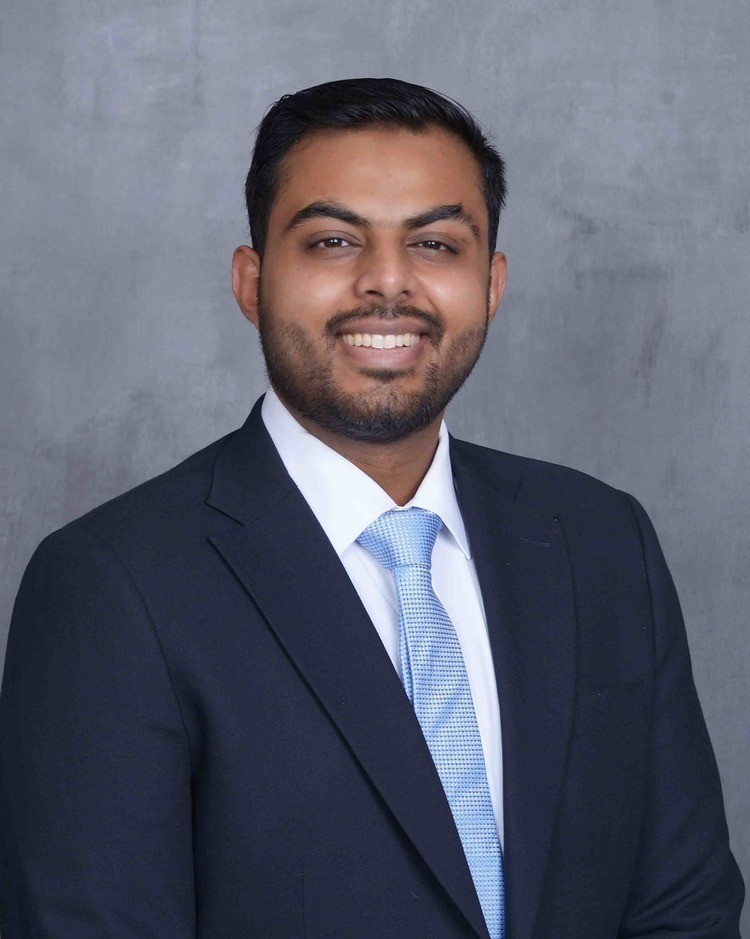 A headshot of Dr. Sajid wearing a navy suit, white button down shirt, and blue tie.