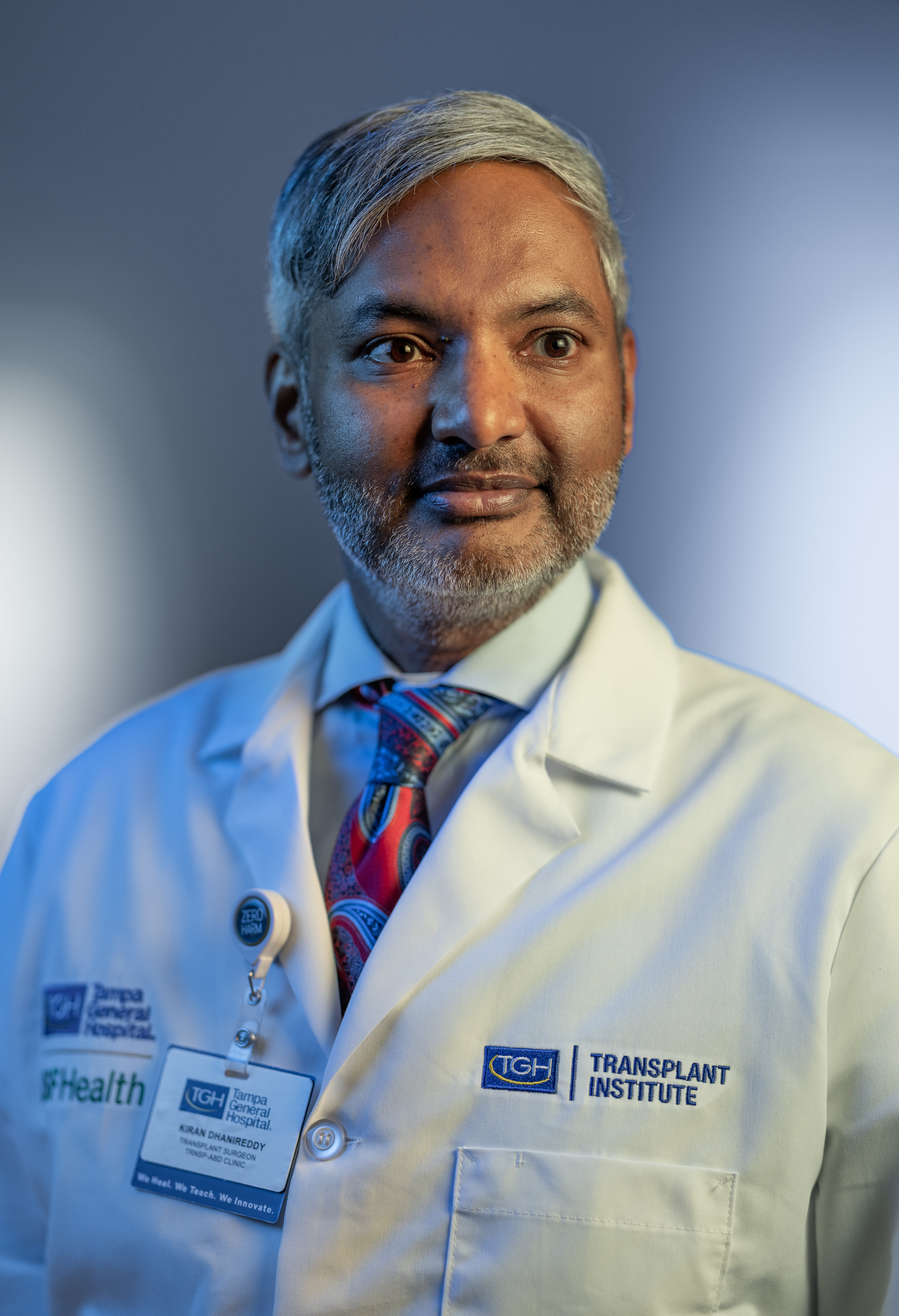 Dr. Kiran Dhanireddy smiling in a medical white coat with a red tie and blue shirt