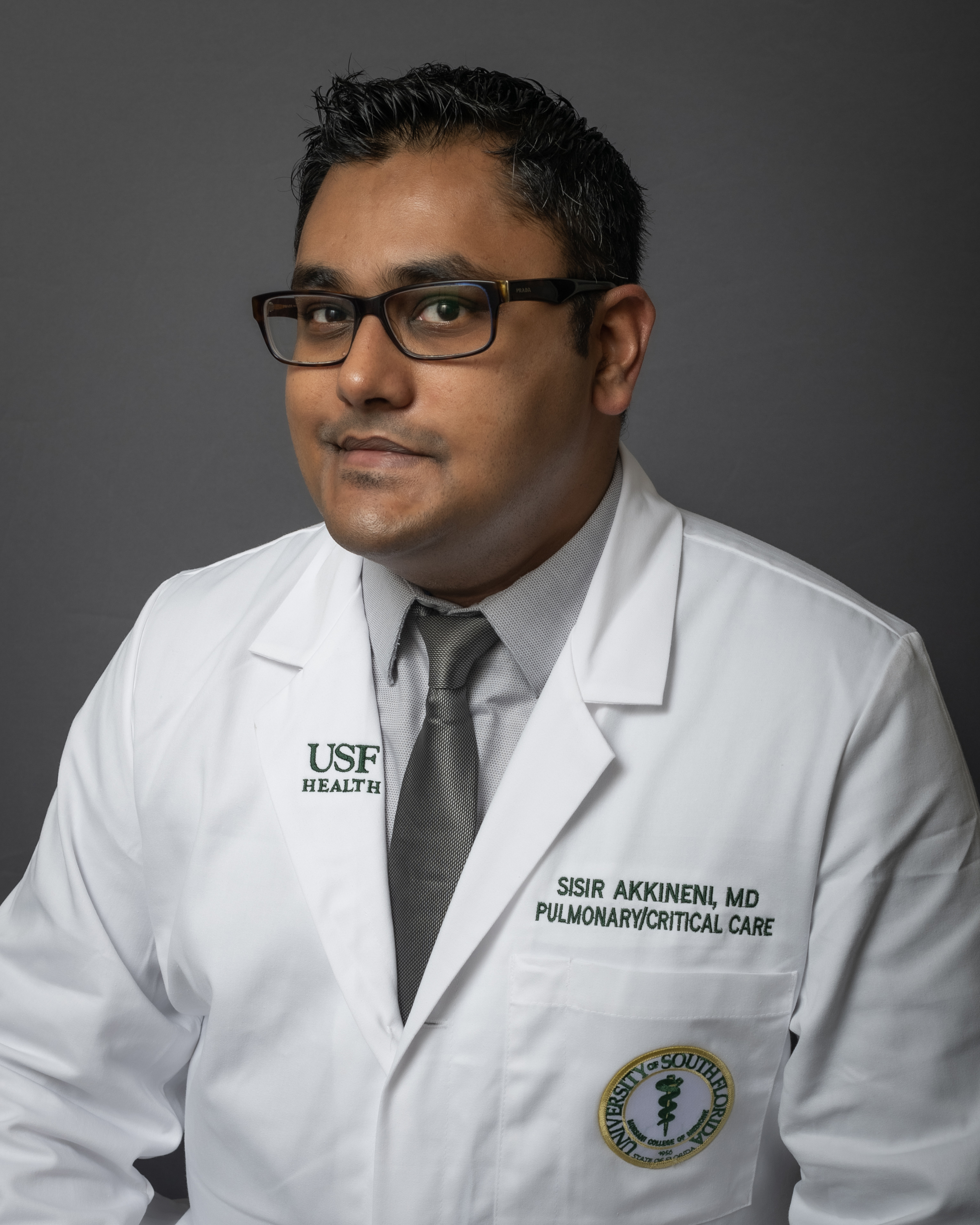 Dr. Sisur Akkineni smiling in a medical white coat with a grey shirt and tie