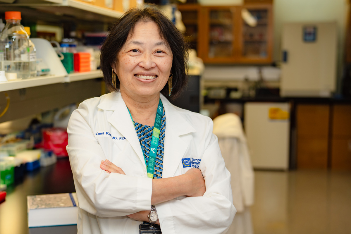 Dr. Kami Kim poses with her arms crossed wearing a medical white coat standing in a lab