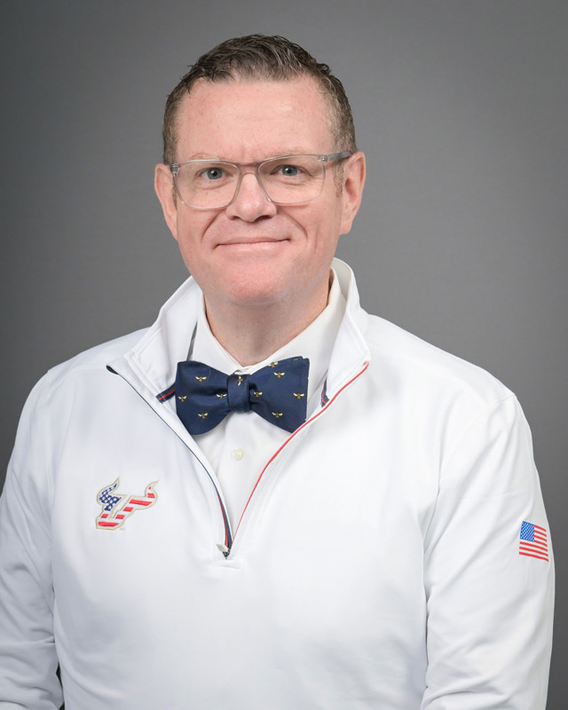 Donald Mullins smiles wearing a white quarter-zip with an American Flag branded USF logo and bow tie with embroidered bees