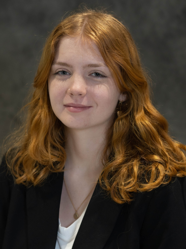 A headshot of Kayleigh Murray smiling in front of a dark grey backdrop. She is wearing a white top and a black blazer.