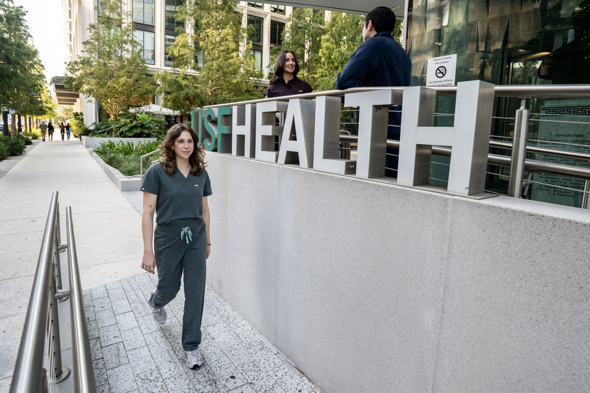 Medial student wearing scrubs walks alongside USF Health letters in front of downtown Tampa building