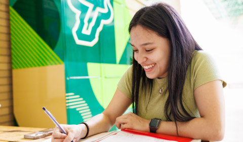 A student writing in a notebook and smiling on campus
