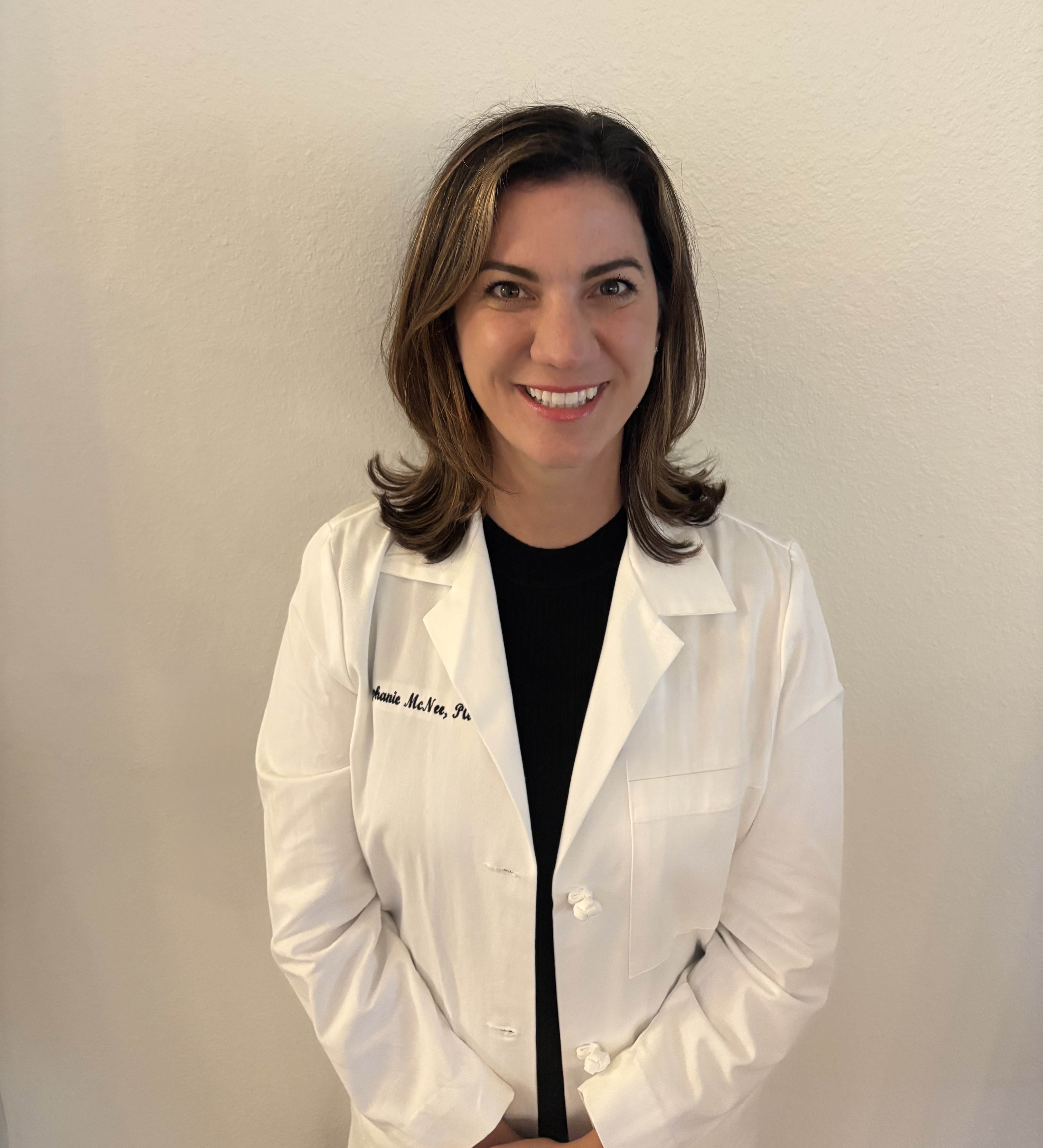 Dr. McNee smiling and standing in front of a white wall wearing a medical white coat wearing black clothing underneath