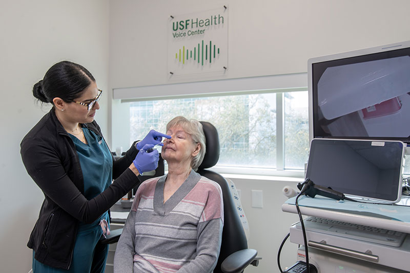 Dr, Abdel-Aty performing a voice dilation on a patient in a medical room, USF Health Voice Center logo is seen on the wall
