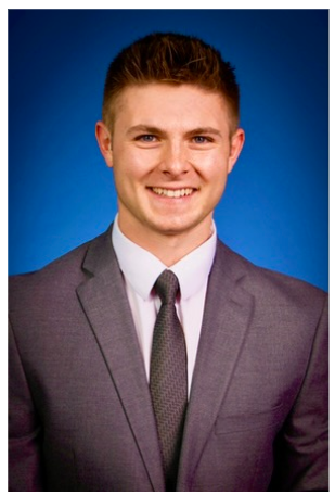 Tristan tinny smiling for a headshot wearing a grey suit and tie with a royal blue background