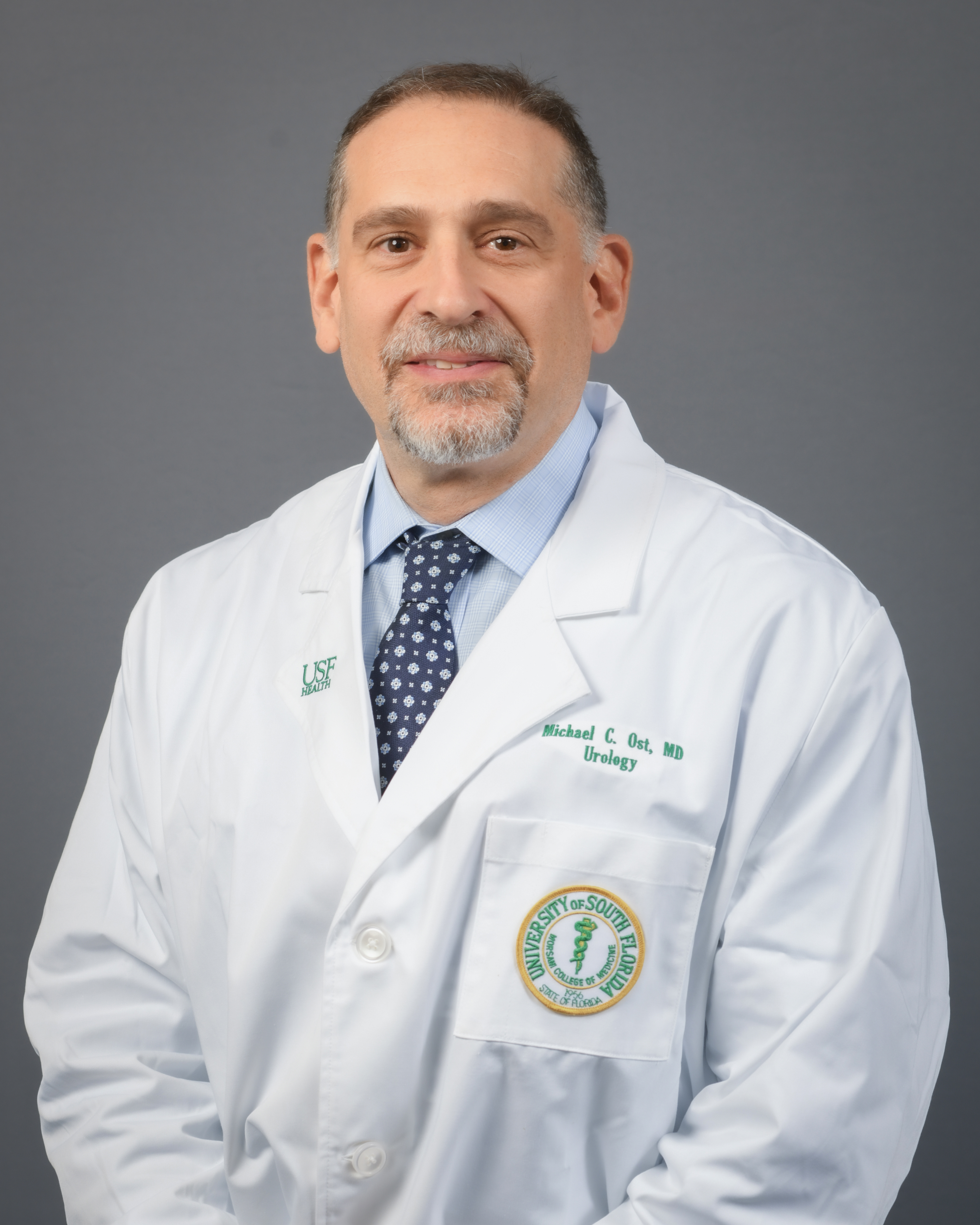 Dr. Ost smiles for a headshot photo wearing a USF Health branded medical white coat with a blue dress shirt and navy blue tie