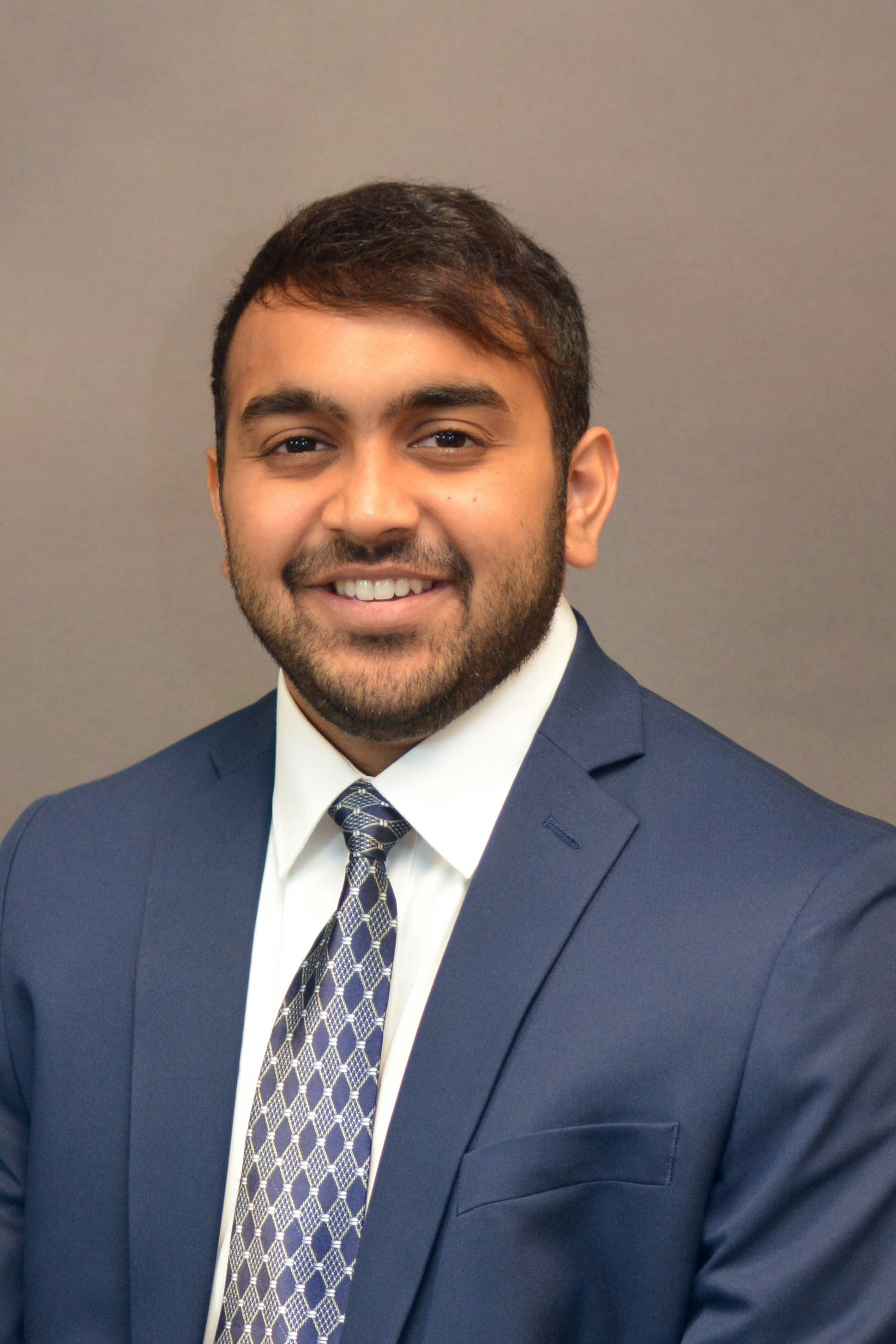 Vikhram Balagee smiling for a headshot wearing a blue suit and tie in a brown background