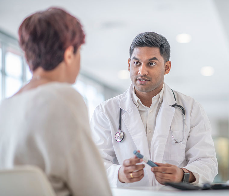 A female adult is at a check up appointment for a diabetic medical condition. She is sitting across the table from her Indian doctor who is holding a glucometer pen that is used to test her blood sugar levels.