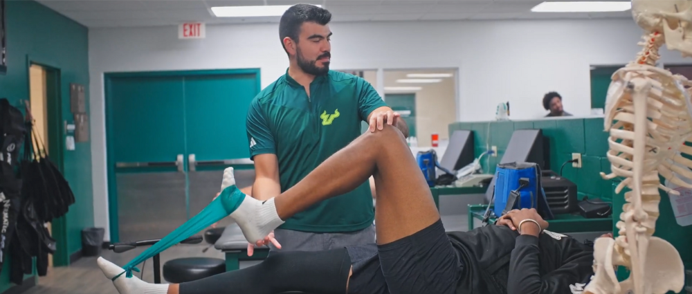 A physical therapist supports a patient’s leg during a resistance band exercise in a rehab clinic, with treatment tables and medical equipment in the background.