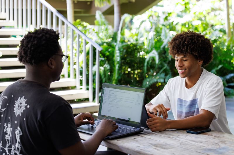 Two men sitting at a table outside. The man on the left is typing on a computer. Both are smiling.