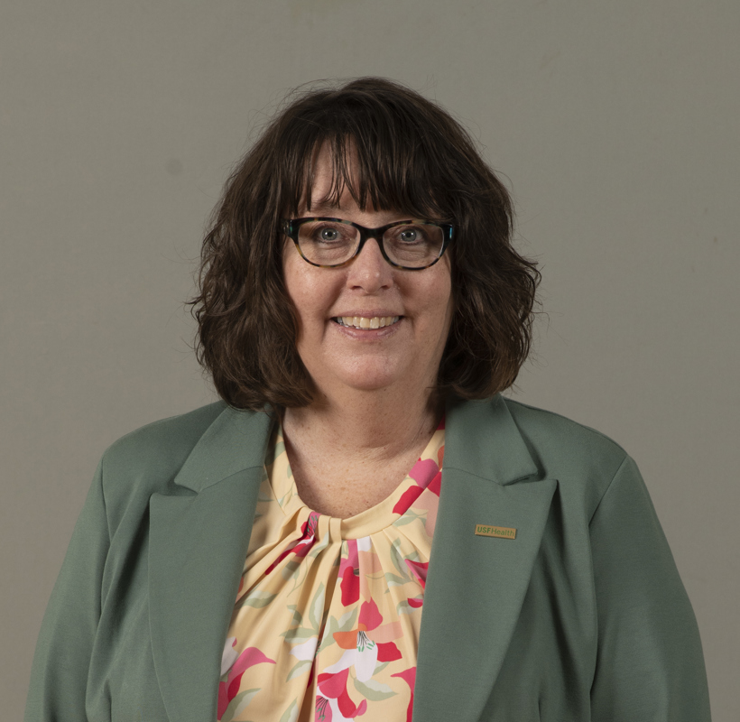 Kathleen Curp smiling wearing a sage blazer and floral blouse in front of a grey backdrop