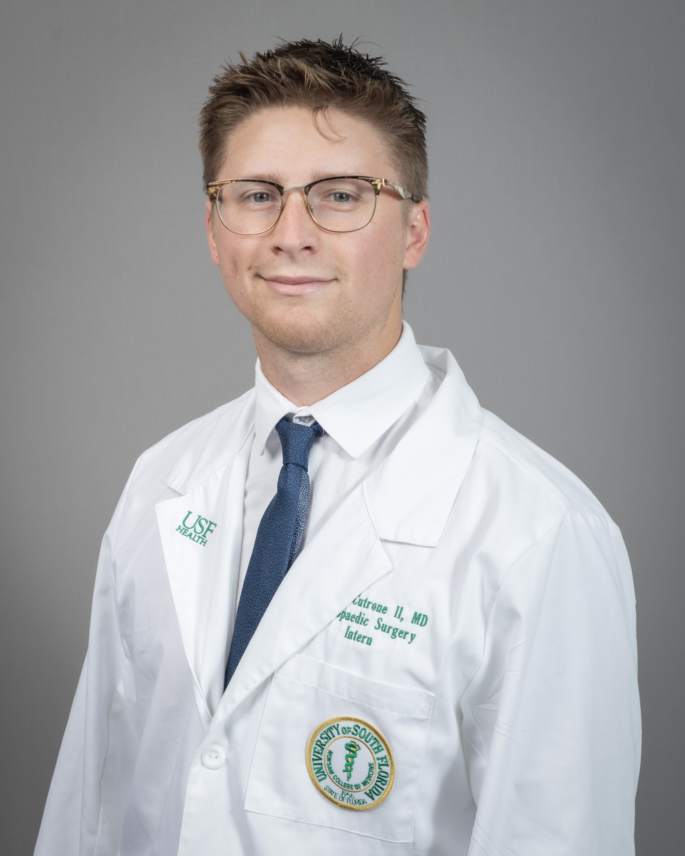 Brock smiling wearing a white dress shirt and blue tie wearing a USF Health branded medical white coat