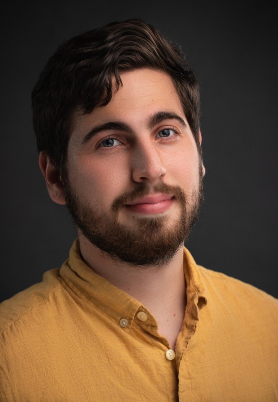 Ryan Rossy posing wearing a mustard yellow button down shirt in front of a black backdrop