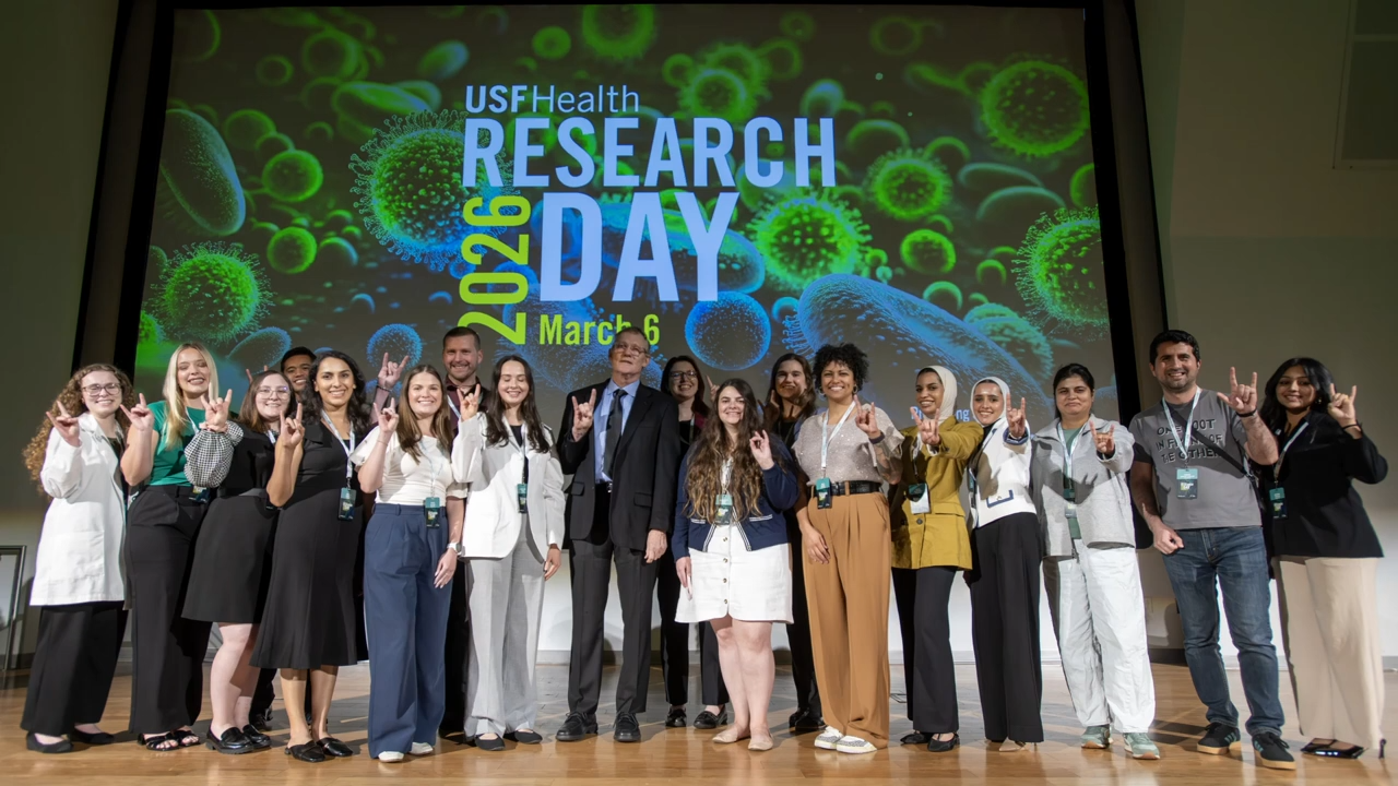 A group of students holding up the "Go Bulls!" in front of a poster that reads "Research Day 2026"