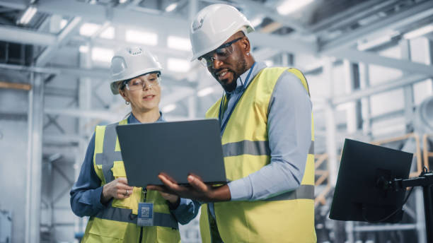 Two workers wearing safety helmets, goggles, and vests in a warehouse looking at a computer