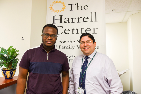 Two men stand in front of a banner displaying "The Harrell Center," smiling and engaged in conversation.