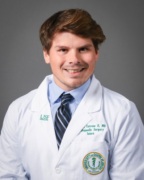 Joseph smiling wearing a blue dress shirt and tie wearing a USF Health branded medical white coat