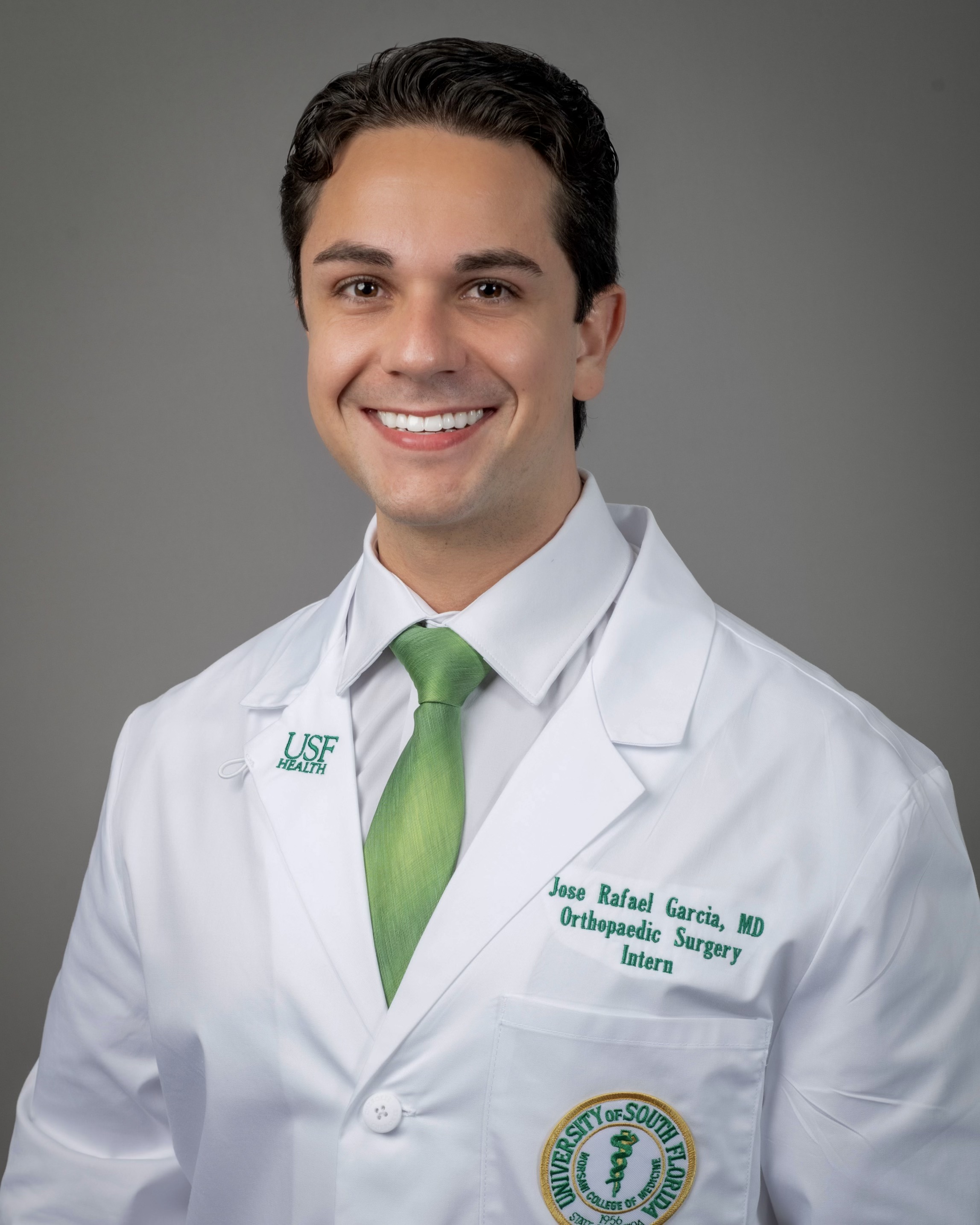 Jose smiling wearing a white dress shirt and green tie wearing a USF Health branded medical white coat