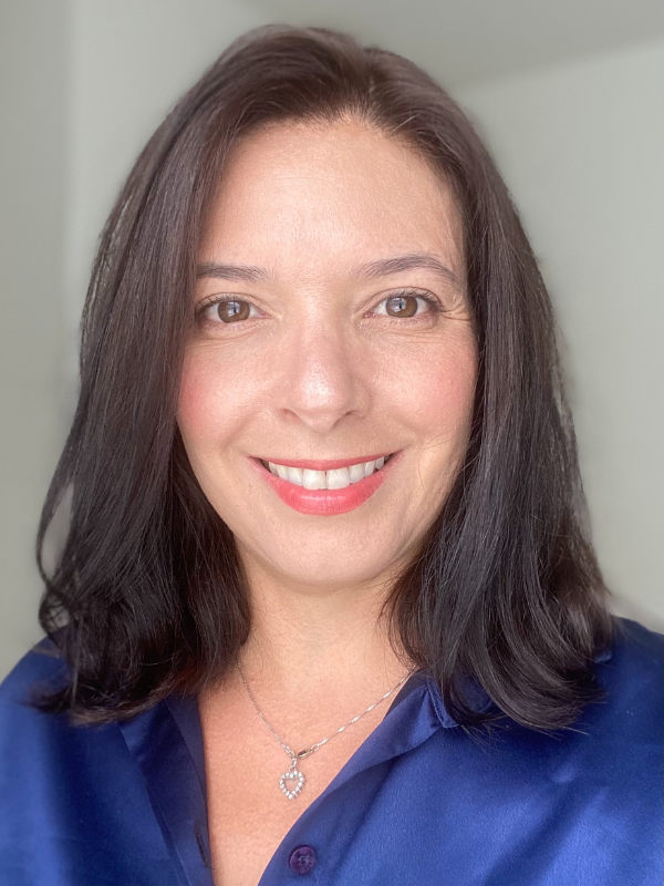 A headshot of Beatriz Rocha smiling in front of a light grey backdrop. She is wearing a blue button-up top.