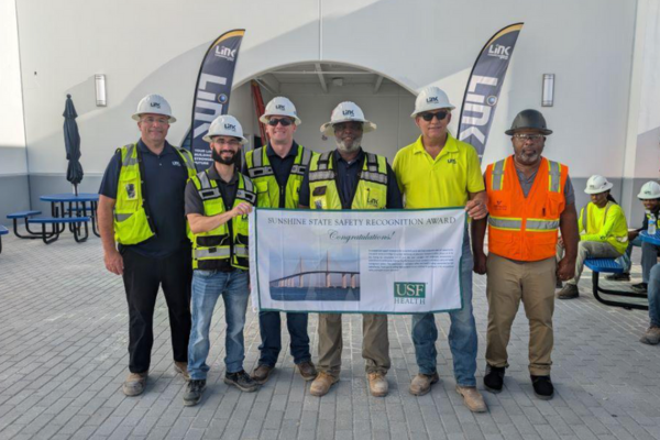A group of construction workers posing for a picture with a USF Health banner.