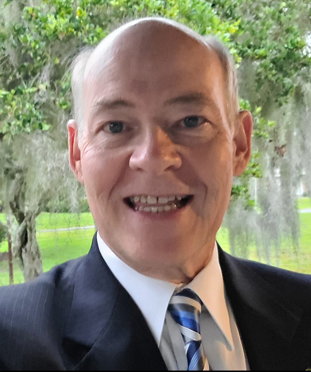 Dr. Goodwin smiling wearing a navy blue suit with white dress shirt and blue striped tie, landscape background